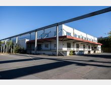 Industriegebäude mit flachem, rechteckigem Grundriss, weißen Fassaden und einer roten Vordachkonstruktion über mehreren Verladerampen. Vor dem Gebäude befindet sich eine asphaltierte Fläche, im Hintergrund Bäume und ein blauer Himmel.