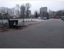 Ein leerer Parkplatz mit gepflastertem Boden, auf dem einige Autos geparkt sind. Im Hintergrund sind kahle Bäume, Bauzäune und moderne Bürogebäude zu sehen, während im Vordergrund eine Mülltonne und eine Schubkarre stehen.