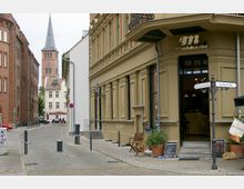 Gepflasterte Straßen einer Altstadt mit historischen Backstein- und Putzfassaden, rechts ein Café mit Außenbestuhlung und ein Straßenschild für die Böttcherstraße und Grünstraße. Im Hintergrund eine Kirche mit hohem Turm und spitzer Turmspitze.