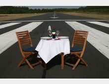 Ein kleines Tischset mit weißer Tischdecke, zwei hölzernen Klappstühlen und gedecktem Geschirr steht auf einer Landebahn mit weißen Markierungen, umgeben von Grasflächen und Wäldern im Hintergrund.