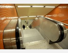 Eine Rolltreppe führt in eine U-Bahn-Station mit orangefarbenen Wandfliesen und metallischen Handläufen. Im Hintergrund ist eine Person auf einer Treppe zu erkennen.