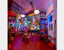 Innenraum eines bunt beleuchteten Cafés oder einer Bar in einem industriellen Gewölberaum mit sichtbaren Leitungen, Hängelampen und zahlreichen Neonbuchstaben an den Wänden. Im Raum stehen kleine Tische und Stühle, Pflanzen sowie metallene Regale mit Leuchtschildern.