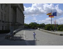 Teilansicht eines historischen Gebäudes mit Säulenfassade (vermutlich der Reichstag in Berlin), flankiert von einer gepflasterten Rampe. Rechts wehen Fahnen, darunter die deutsche Flagge, die EU-Flagge und weitere, im Hintergrund sind Bäume und Personen zu sehen.