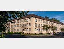 Eckhaus aus beigefarbenem Putz mit drei Stockwerken und zahlreichen Fenstern, typisch für ältere Wohngebäude; davor eine Straßenecke mit Gehwegen, Straßenlaternen, Verkehrszeichen und vereinzelten Bäumen. Im Vordergrund parkende Autos und Teile eines Straßenschildes sichtbar.