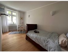 Schlichtes Schlafzimmer mit Holzboden, einem Einzelbett aus dunklem Holz auf der rechten Seite und einem kleinen weißen Schreibtisch mit Stuhl vor einem Fenster mit weißen Vorhängen. Ein gestreifter Teppich liegt neben dem Bett.