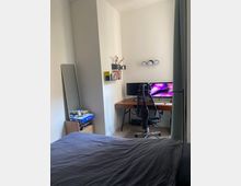 Kompaktes Schlafzimmer mit einem Schreibtisch und Bürostuhl in einer Ecke. Auf dem Schreibtisch stehen zwei Monitore, darüber kleine Wandregale mit Büchern und Deko, daneben ein hoher Spiegel und ein Regal mit Büchern und Gegenständen.