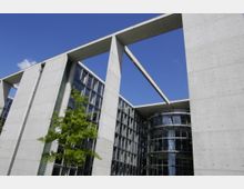 Modernes Bürogebäude aus Beton mit großen Glasfassaden und einem auffälligen rechteckigen Rahmen. Im Vordergrund ist ein grüner Baum zu sehen, der einen Kontrast zum klaren blauen Himmel bildet.