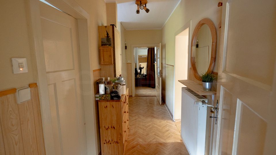 Schmaler Wohnungsflur mit hellem Fischgrät-Parkett, beige gestrichenen Wänden und mehreren weißen Türen entlang des Gangs. An der Wand stehen eine Holzkommode und ein Wandspiegel über einem kleinen Regal neben einem Heizkörper; am Ende führt eine geöffnete Tür in einen weiteren Raum.