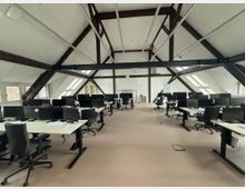 Großraumbüro unter einem Dachstuhl mit sichtbaren Holzbalken, zahlreichen Schreibtischen mit Bürostühlen und Computermonitoren. Tageslicht strömt durch schräge Dachfenster und rechteckige Fenster an den Seiten.