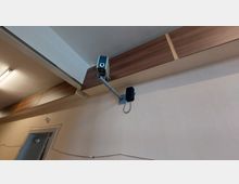 Überwachungskamera, die an einer beige gestrichenen Wand montiert ist, mit einer Halterung und sichtbarem Kabel. Im Hintergrund sind Holzelemente an der Decke sowie eine Tür und eine Deckenleuchte zu erkennen.