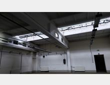 Industriehalle mit betonierten Wänden und Decke, mehreren Oberlichtern für Tageslicht sowie hängenden Leuchtstofflampen. An den Wänden sind Rohrleitungen und Heizkörper befestigt, eine Tür führt in einen angrenzenden Raum.