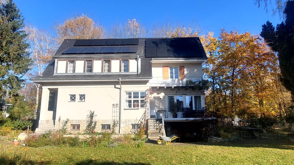 Freistehendes zweigeschossiges Wohnhaus mit hellem Putz, dunklem Mansarddach und mehreren Dachgauben; auf dem Dach sind Solarpaneele montiert. Vor dem Haus liegen ein Garten mit Rasenfläche sowie Bäume mit Herbstlaub rundherum.