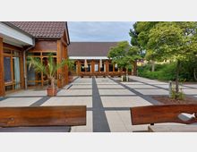 Moderner Innenhof mit gepflastertem Bodenmuster, das aus hellen und dunklen Fliesen besteht. Umgeben von einem Holzgebäude mit großen Fenstern, befinden sich im Hof dekorative Palmen in Töpfen, Sitzbänke und Begrünung mit Bäumen.