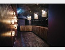 Moderner Garderobenbereich mit hölzerner Theke, dekorativer Wandbeleuchtung und hängenden Deckenlampen. Die Decke ist dunkel und teilweise gitterförmig, der Raum erscheint gedämpft und elegant beleuchtet.