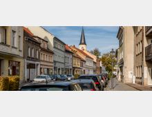 Straßenszene mit dicht geparkten Autos und einer Reihe von zweistöckigen Wohn- und Geschäftshäusern unterschiedlicher Fassadenfarben; im Hintergrund ein Kirchturm mit spitzem Dach, flankiert von Bäumen.