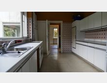 Küche mit weißen Einbauschränken und Arbeitsflächen auf beiden Seiten. Die Wände sind teils mit orangefarbenen, gemusterten Kacheln verkleidet, und ein Fenster bietet Ausblick in einen Gartenbereich. Eine Tür im Hintergrund führt zu einem angrenzenden Raum.