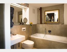 Modern eingerichtetes Badezimmer mit beiger Wandverkleidung aus Fliesen, einer weißen Badewanne und einem rechteckigen Waschbecken. Im Raum befinden sich ein großer goldgerahmter Spiegel, dekorative Objekte und ein an die Wand gelehntes Stab-Element mit einer Sonnenblume.