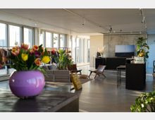 Heller, modern eingerichteter Innenraum mit großen Panoramafenstern, die viel Tageslicht einlassen. Im Vordergrund befindet sich eine violette Vase mit bunten Tulpen, während der Raum mit Sitzmöbeln, Pflanzen und Kleiderständern ausgestattet ist.