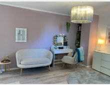 Helles Wohnzimmer mit rosafarbenen Wänden und grauem Laminatboden, eingerichtet mit einem cremefarbenen Sofa, einem Schminktisch mit Spiegel und passendem Sessel sowie einem runden Teppich. Rechts stehen ein Sideboard mit Stehlampe und ein Regal in einer Nische, darüber hängt eine große Deckenleuchte mit Stoffschirm.