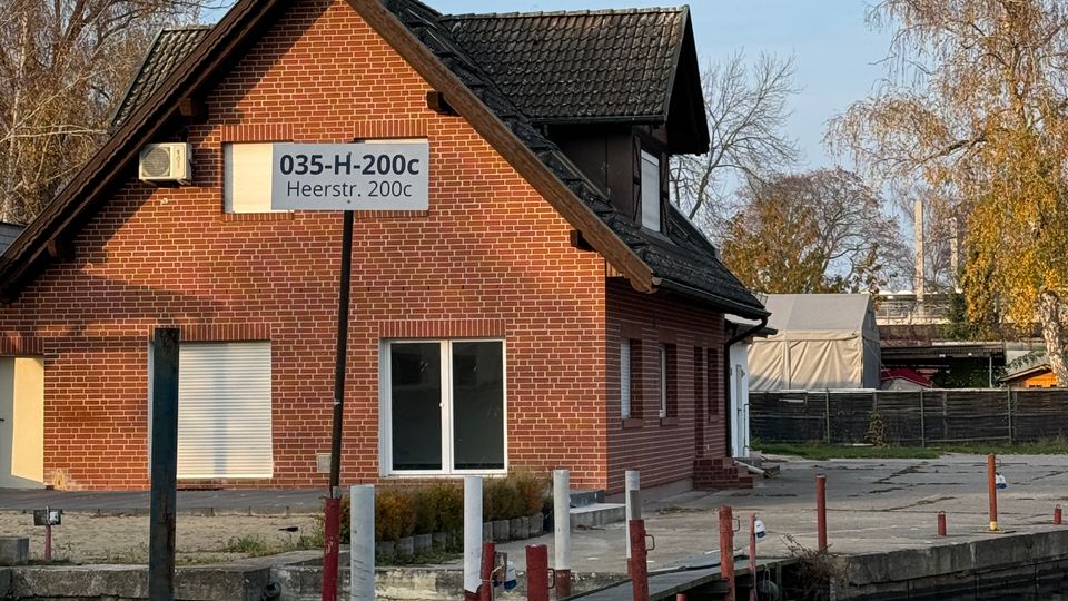 Backsteinbau mit Satteldach am Ufer eines Gewässers, davor ein kleiner Steg mit Pfosten. Im Hintergrund sind Bäume und ein umzäuntes Gelände sichtbar.