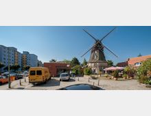 Historische Windmühle mit großen Flügeln, umgeben von einem gepflasterten Platz mit geparkten Autos, einem kleinen roten Gebäude mit der Aufschrift "Haar Design" und einem Cafébereich mit Sonnenschirmen; im Hintergrund moderne Wohngebäude und Bäume.