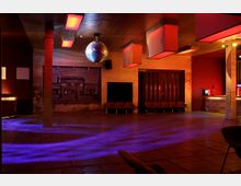 Ein leerer, moderner Veranstaltungsraum mit Holzboden und indirekter Beleuchtung in roten und violetten Tönen. Eine Discokugel hängt an der Decke, an den Wänden befinden sich Bilder, und eine Sitzbank ist an der Rückwand platziert.