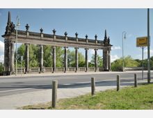 Historische Steinarkade mit mehreren Säulen und dekorativen Verzierungen, flankiert von zwei Obelisken; im Vordergrund eine Straße mit einem gelben Richtungsschild nach Berlin und Potsdam sowie einem Bürgersteig. Im Hintergrund sind Bäume und ein klarer Himmel sichtbar.