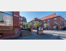 Straßenecke mit roten Backsteingebäuden im klassischen Stil, umgeben von kleinen Bäumen und Gehwegen. Im Vordergrund befinden sich eine Treppe, ein Freistehender Schaukasten aus Backstein mit Glasdach sowie ein Straßenschild mit der Aufschrift „August-Bebel-Straße“.