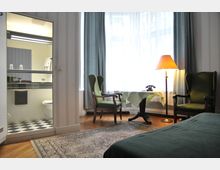 Ein gemütliches Hotelzimmer mit grün gepolsterten Sesseln, einem kleinen runden Tisch mit Tischdecke, einer Stehlampe und einem schwarzen Telefon. Im Hintergrund ein Badezimmer mit schwarz-weiß kariertem Boden und einem Waschbecken, das durch eine offene Tür sichtbar ist.