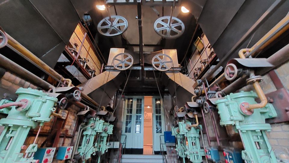 Industrieller Maschinenraum mit alten mechanischen Vorrichtungen, darunter große Zahnräder, Rohre und grün lackierte Maschinen. Der Raum hat eine Backsteinwand und eine doppelflügelige Tür im Hintergrund.