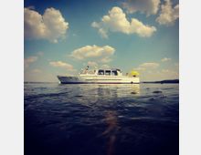 Ein weißes Motorboot liegt auf ruhigem Wasser, umgeben von einer weitläufigen Seelandschaft unter einem Himmel mit vereinzelten Wolken. Auf dem Boot sind Menschen zu sehen, darunter eine Person unter einem gelben Sonnenschirm.