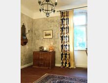 Ein gemütlicher Raum mit rustikaler Einrichtung: alte, rau verputzte Wände, eine Holzkiste mit drei Schubladen, darauf eine Kerze und Dekoration. Rechts ein großes Fenster mit gemusterten Vorhängen, oben ein metallener Kronleuchter, am Boden ein orientalischer Teppich.