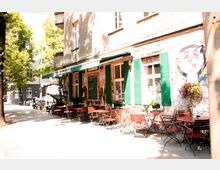 Eine Straßenszene mit einer kleinen Caféterrasse vor einem historischen Gebäude mit rauen Backsteinmauern und grünen Fensterläden. Die Terrasse ist mit Holzstühlen und runden Tischen ausgestattet, umgeben von Bäumen und dekoriert mit Blumen.