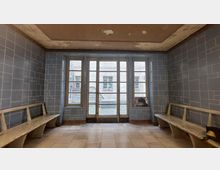 Ein leerer, alter Raum mit blauen Wandfliesen, zwei Holzbänken an den Seiten und einer großen Fensterfront aus mehreren Glaselementen in der Mitte. Die Fenster bieten einen Blick auf einen Innenhof mit weißen Ziegelwänden.