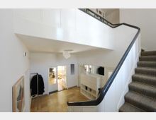 Eingangsbereich eines Hauses mit Treppenaufgang, weiß gestrichenen Wänden und einem Flur mit gelblichen Bodenfliesen. Im Hintergrund befindet sich eine Doppeltür mit Glaseinsätzen, die einen Blick in einen hell erleuchteten Raum mit Stühlen ermöglicht.