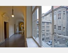 Ein langer Gang mit glänzendem Boden, verzierten Türen und Bogenwänden auf der linken Seite sowie großen Fenstern rechts, die den Blick auf die Fassade eines historischen Gebäudes mit Steinverzierungen und Sprossenfenstern freigeben.