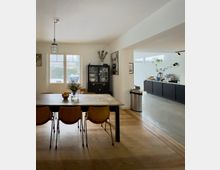 Offener Wohn- und Essbereich mit großem Holztisch und Stühlen vor einer Glastür zum Garten, daneben ein dunkler Vitrinenschrank. Rechts schließt sich eine moderne Küche mit langen, schwarzen Unterschränken, hoher Fensterzeile und Sichtbetonboden an.