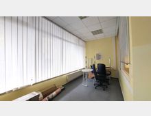 Ein kleiner Büroraum mit gelben Wänden und großen vertikalen Jalousien vor den Fenstern. Der Raum enthält zwei Bürostühle, einen weißen runden Tisch, ein Regal und einige liegende Gegenstände wie Verpackungen am Boden.