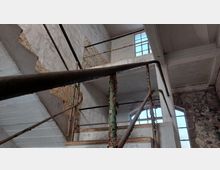 Industriegebäude-Innenraum mit einem verwitterten und teils rostigen Treppengeländer aus Metall. Die Wände sind teils verputzt, teils freiliegend, mit abblätternder Farbe, und große Fenster lassen diffuses Licht einfallen.