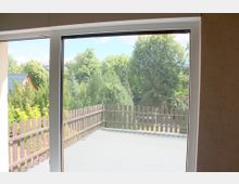 Blick durch ein großes Fenster auf eine Terrasse mit Holzzaun, dahinter Bäume und Vegetation; im Hintergrund ist ein Haus mit dunklem Dach erkennbar.