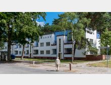 Wohngebäude im Bauhausstil mit heller Fassade, roten Backsteinelementen und blauen Akzenten, umgeben von Bäumen und Grünflächen. Im Vordergrund eine Straße mit Bürgersteig, auf dem eine ältere Person spaziert, sowie ein Straßenschild mit der Aufschrift „Riemeisterstraße“.