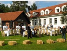 Eine Gartenveranstaltung vor einem historischen Gebäude mit roten Ziegeldächern und weißen Fassaden. Im Vordergrund stehen dekorierte Tische mit weißen Tischdecken, Strohballen auf einer grünen Wiese und mehrere Personen in eleganter Kleidung.
