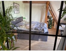 Schlafzimmer in einer Dachwohnung mit sichtbaren Holzbalken und einem großen Bett mit grauem Kopfteil. Im Vordergrund befinden sich Zimmerpflanzen und eine gläserne Raumtrennwand; rechts ist eine kleine Sitzecke sichtbar.