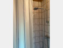 Geflieste Duschkabine mit weißen Wandfliesen und dunklen Fugen, ausgestattet mit Handbrause an einer Stange und Mischarmatur. Links hängt ein weißer Duschvorhang mit zwei blauen Längsstreifen.