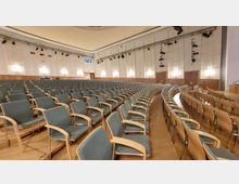 Großer Veranstaltungssaal mit Reihen von gepolsterten Stühlen mit Holzlehnen, angeordnet auf einem Holzfußboden. Die Wände sind hell gestaltet, mit Wandvertäfelungen und integrierter Beleuchtung, sowie einer technischen Ausstattung aus Scheinwerfern an der Decke.