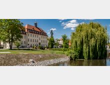 Historisches Gebäude mit hellem Putz und rotem Ziegeldach, flankiert von einem großen Baum und einer Weide, die am Ufer eines kleinen Gewässers steht. Im Vordergrund ist eine Wiese mit Steinböschung zu sehen, während der Himmel blau und wolkig ist.