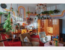 Wohnzimmer mit Retro- und Boho-Elementen, dekoriert mit zahlreichen Zimmerpflanzen, bunten Makramee-Hängern, einer hölzernen Schrankwand und einem Schreibtisch mit Nähmaschine. An der weißen Wand sind bunte Wellenmuster angebracht, und das Zimmer wird von Sonnenlicht durchflutet.