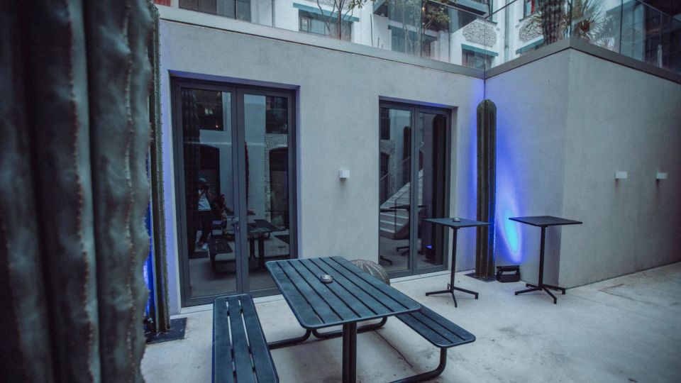 Innenhof-Terrasse eines modernen Gebäudes mit hellen Betonwänden und großen Glas-Schiebetüren. Im Vordergrund stehen ein schwarzer Picknicktisch mit Bänken und zwei Stehtische; hohe Kakteen und blaues Akzentlicht markieren die Ecken.