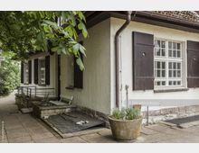 Einfamilienhaus mit weiß verputzter Fassade, dunklen Holzfensterläden und einem kleinen Sitzbereich mit weißer Bank vor dem Eingang. Im Vordergrund stehen Pflanzgefäße, und im Hintergrund sind Gehwege und grüne Bäume sichtbar.