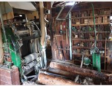 Innenraum einer alten Holzwerkstatt mit einer großen grünen Holzbearbeitungsmaschine im Vordergrund. Im Hintergrund sind Regale mit Werkzeugen, Holzblöcken und weiteren Materialien sowie diverse Rohre und Kabel sichtbar.